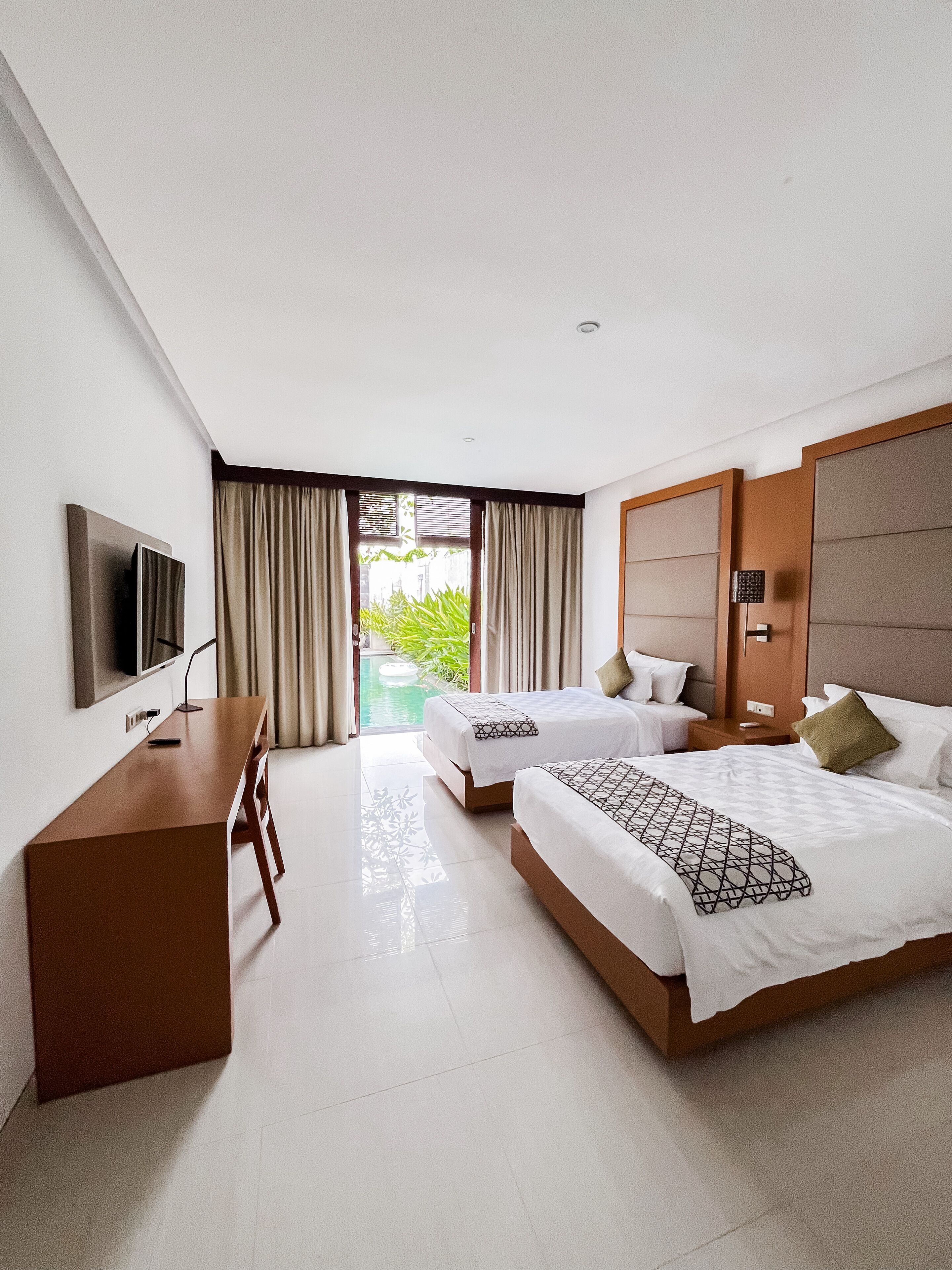 A hotel room with two beds, a desk, a TV, and a view of a swimming pool.