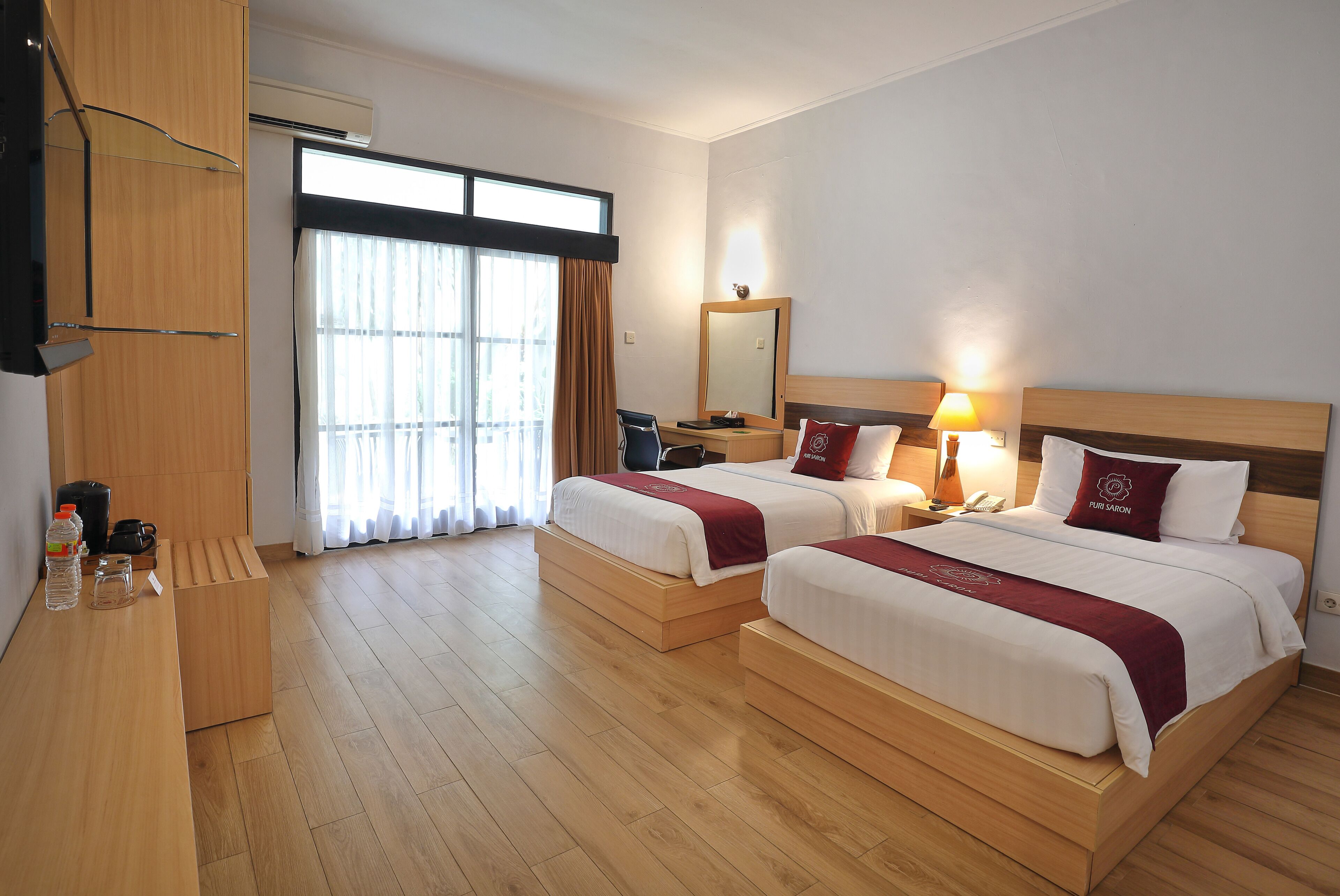 Una camera d'albergo con due letti, una scrivania, una sedia, uno specchio, una lampada e una finestra con tende.