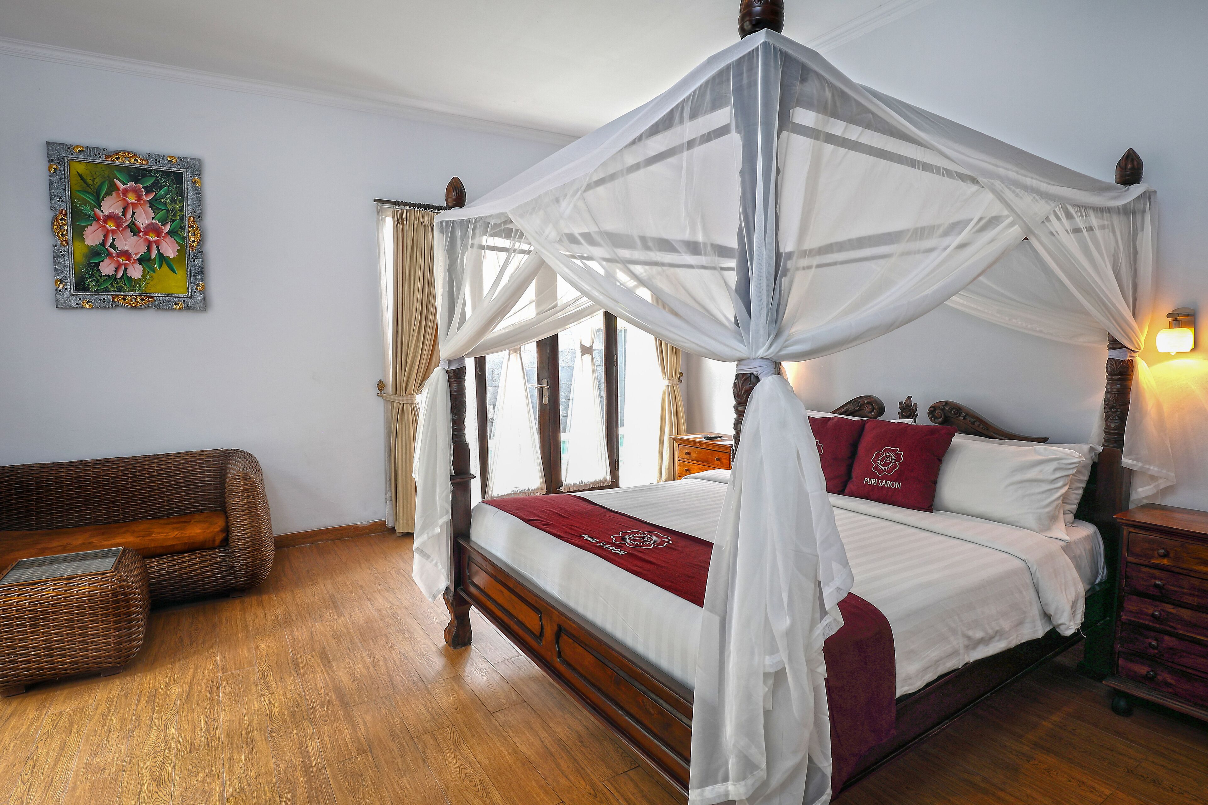 A four-poster canopy bed with a white bedspread and red pillows, a wicker sofa, and a wooden nightstand with a lamp.