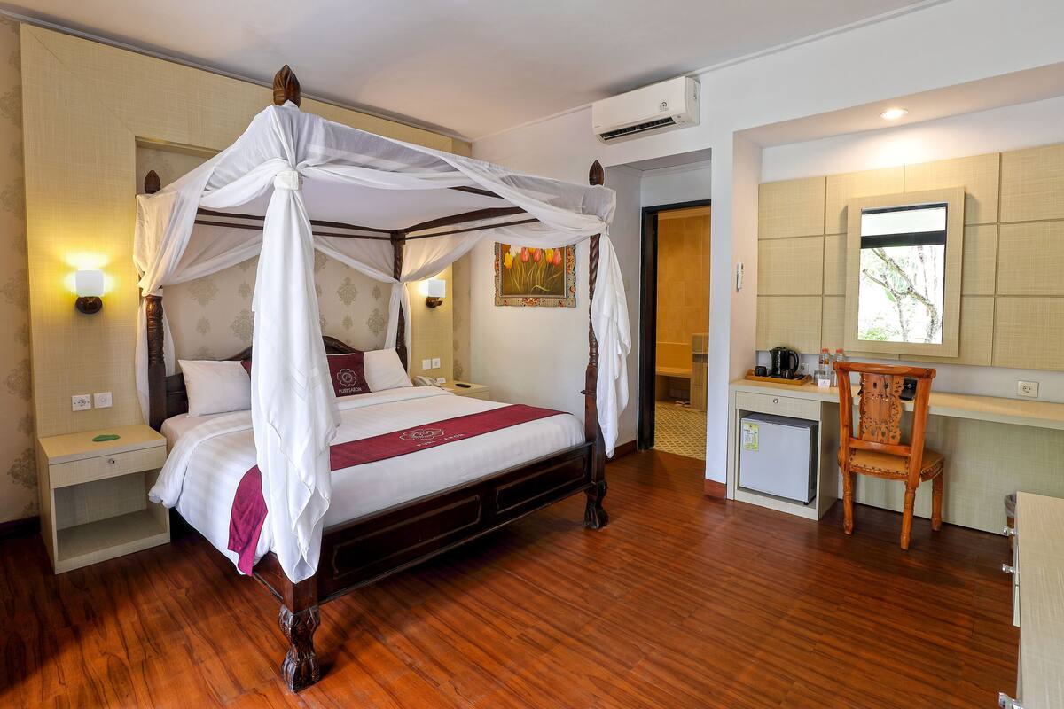 A four-poster bed with a canopy, bedside tables, a small desk with a chair, a refrigerator, and a window with a view.