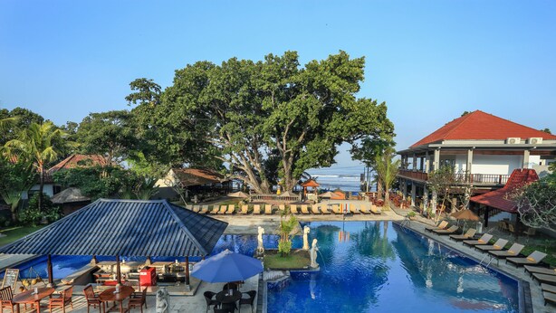 A pool area with lounge chairs and a dining area, surrounded by trees and a building with a red roof. - Puri Saron Seminyak (Seminyak)