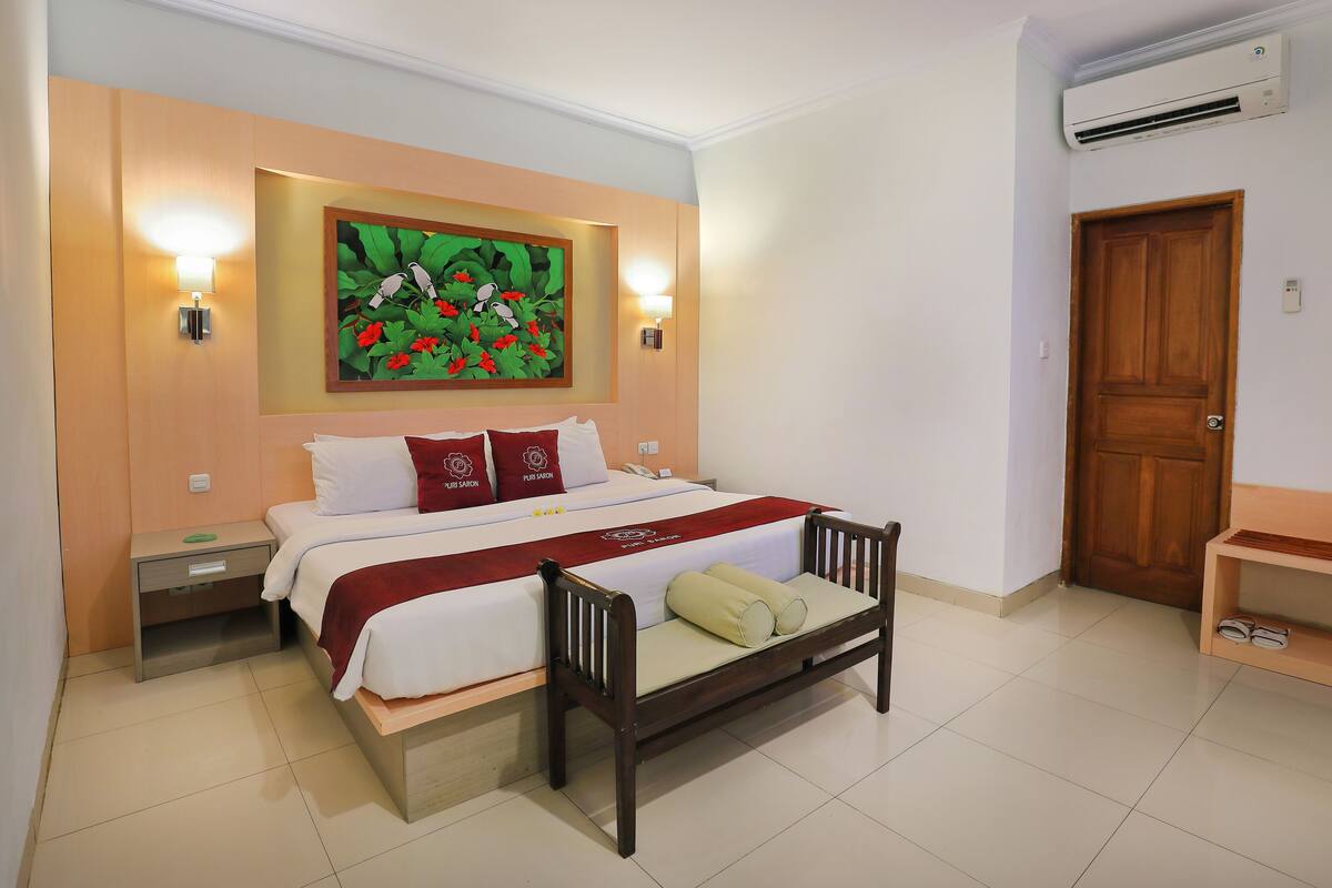 A hotel room with a bed, bedside tables, a bench, a painting, and an air conditioning unit.