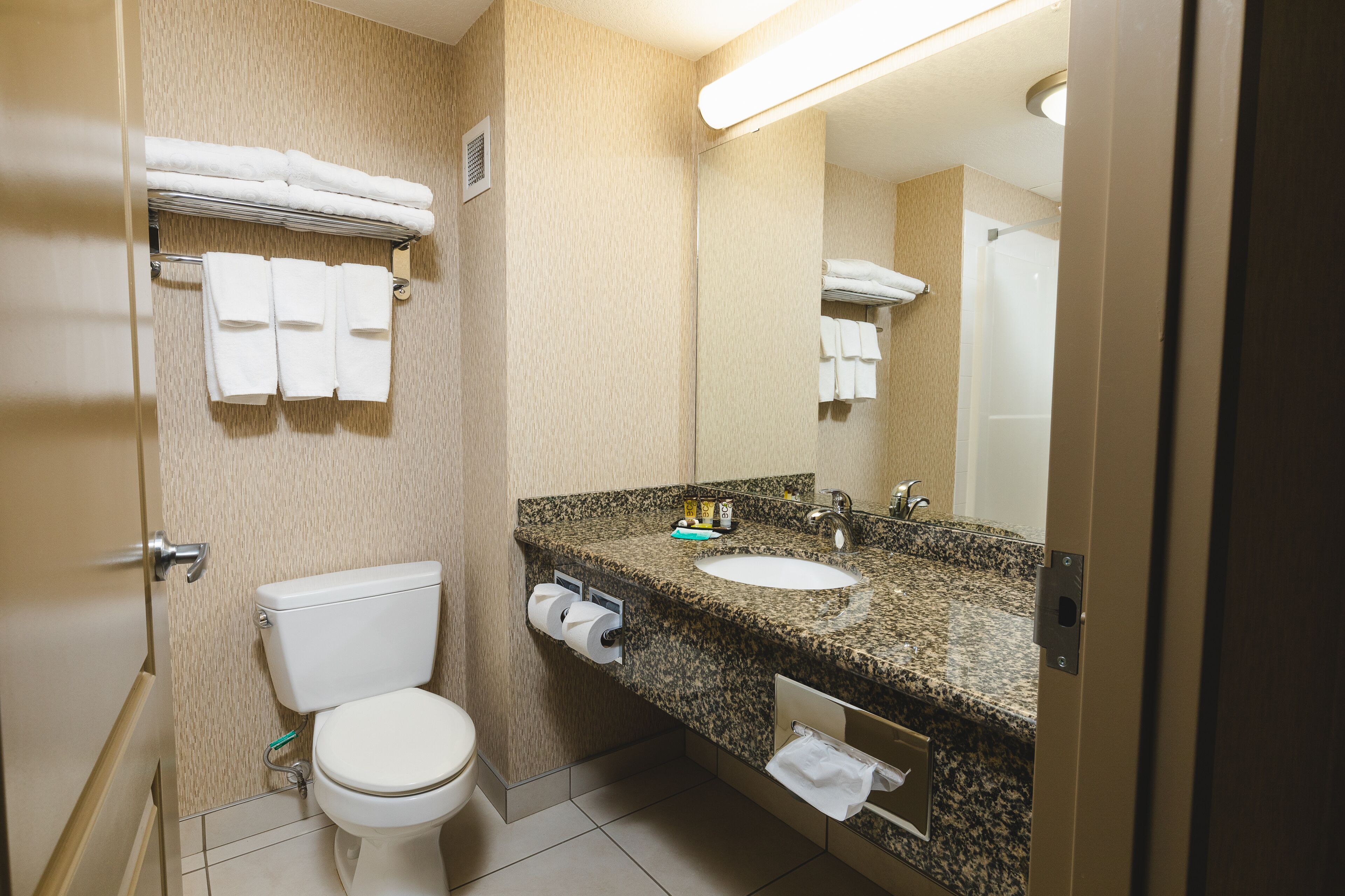 Baño con inodoro, lavamanos y espejo. Hay toallas colgadas en un perchero y un dispensador de toallas.