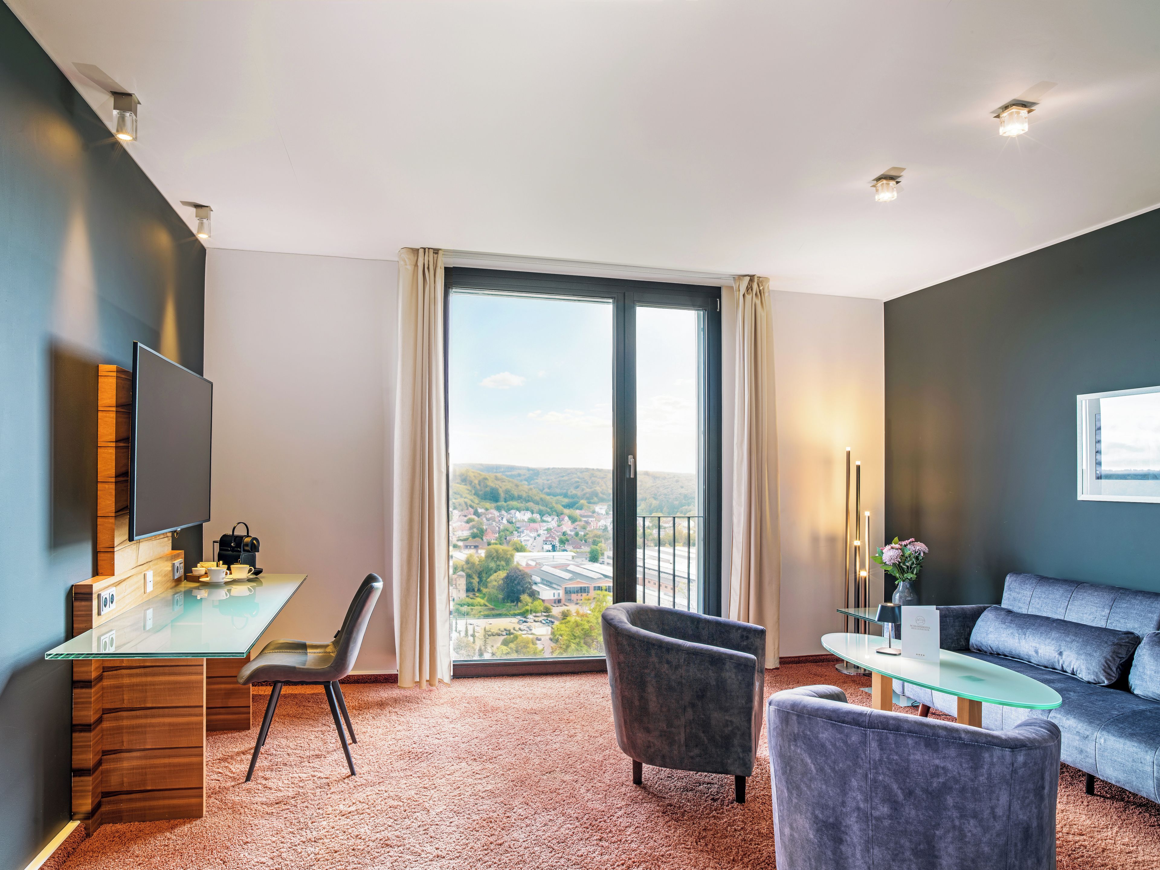 A modern hotel room with a glass desk, a comfortable sofa, and a large window offering a city view.