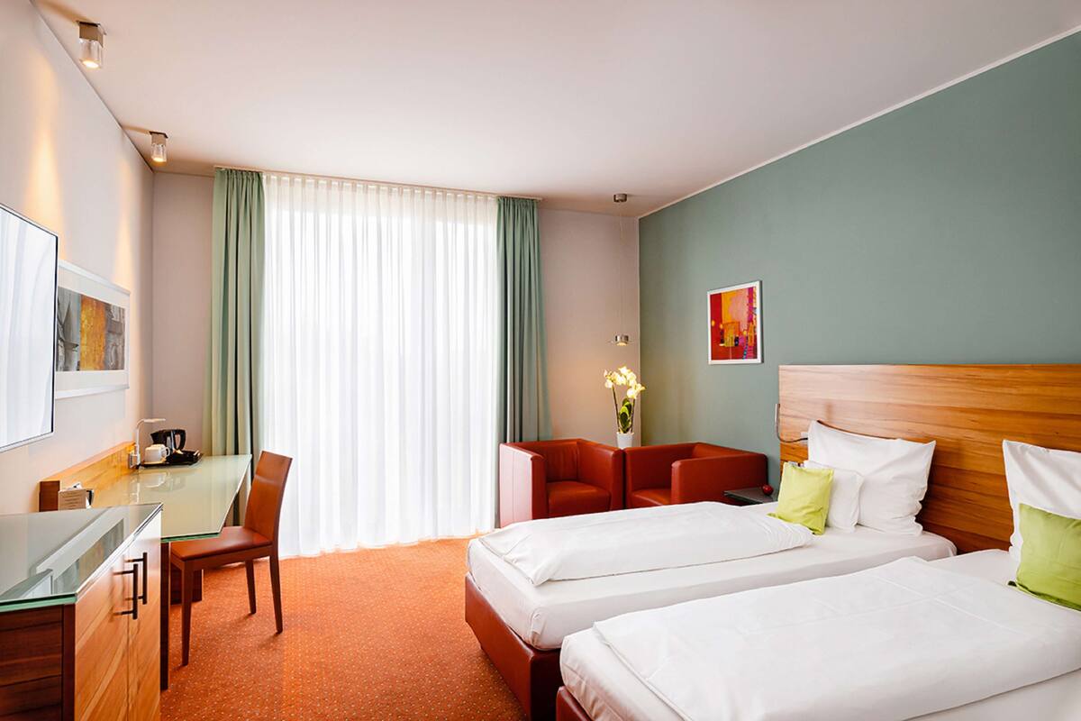 A hotel room with two beds, a desk with a chair, a small table, and a window with curtains.