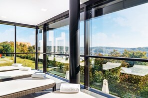 Une salle de spa moderne dotée de grandes fenêtres, offrant une vue sur la campagne, et comprenant plusieurs tables de massage avec des serviettes.