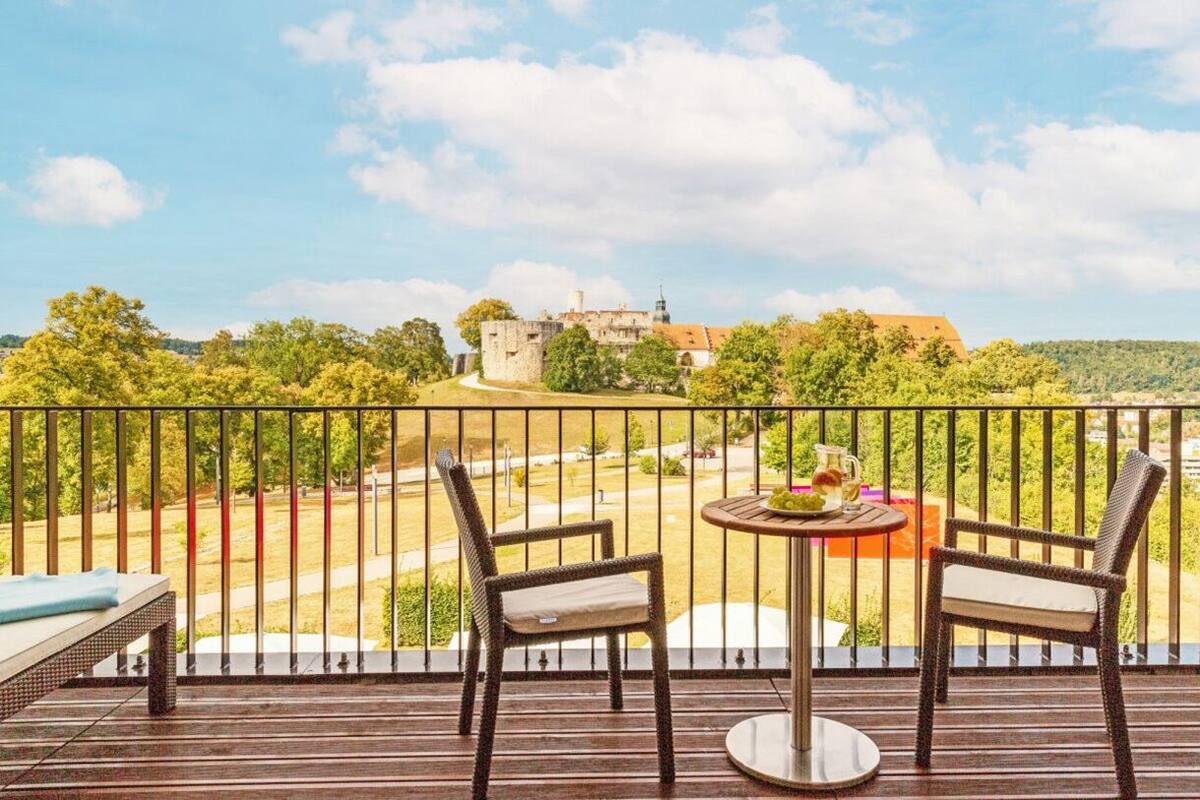 A balcony with two wicker chairs and a small table, overlooking a castle and lush greenery.