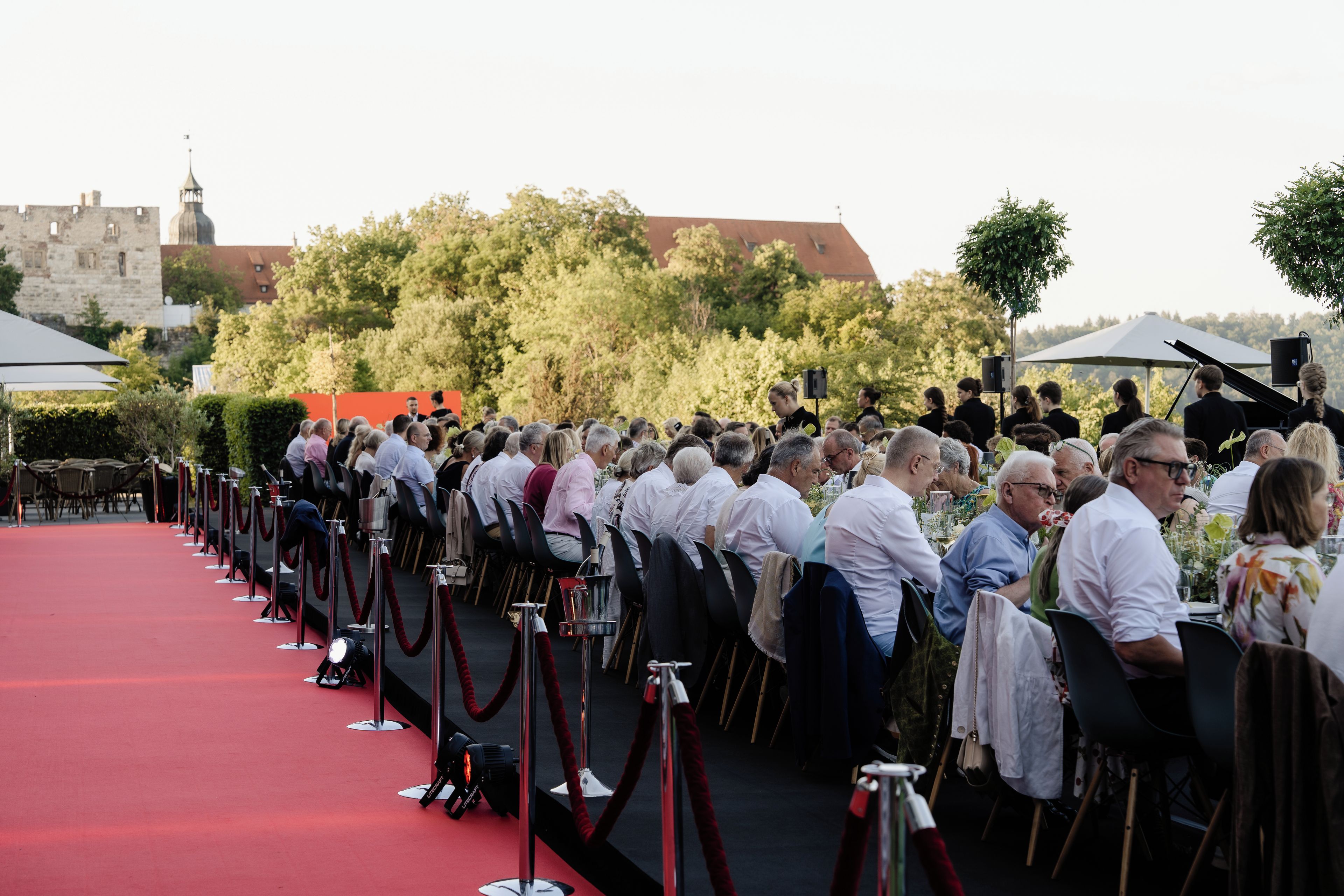 A red carpet event with attendees seated in rows, a stage with musical equipment, and a historic building in the background.