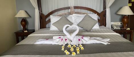 A neatly made bed with a heart-shaped towel arrangement, a floral centerpiece, and a wooden headboard.