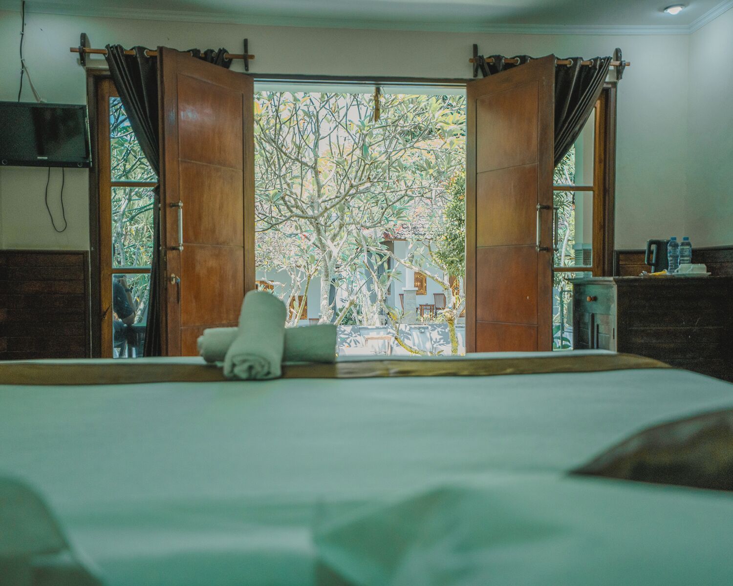 A room with a bed, a window with open curtains, a view of trees, and a small table with a towel.