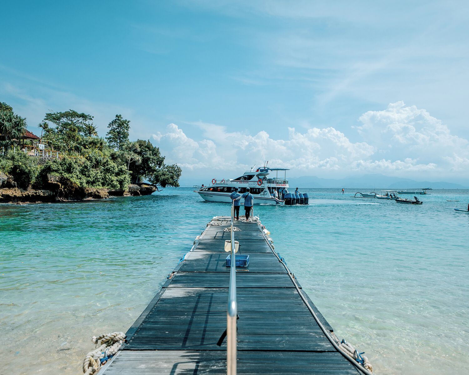A pier extending into clear water with boats and a house on the shore.