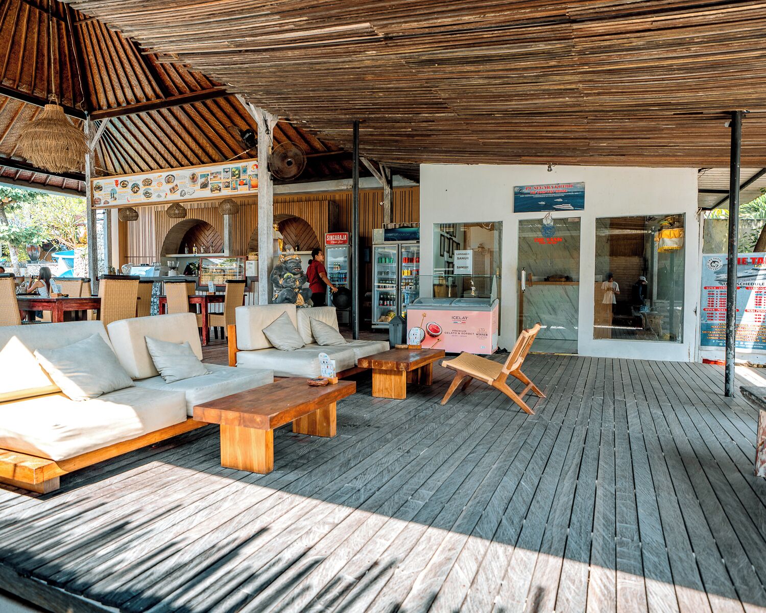 Uma área de estar ao ar livre com móveis de madeira, um espaço coberto com telhado de palha e uma fachada com refrigerador e placas.