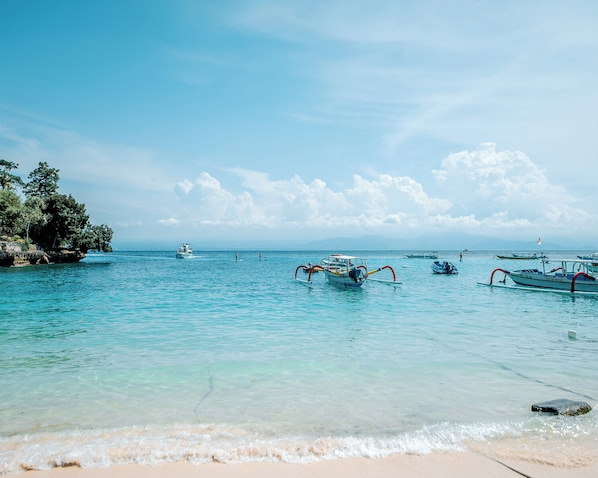 A beach with clear turquoise water, boats, and a rocky outcrop with vegetation.
