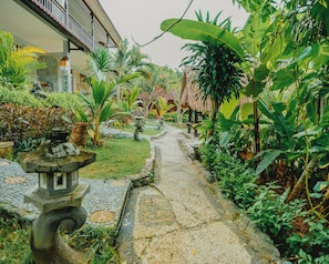 A garden pathway with stone tiles, surrounded by lush tropical plants and a traditional building with a tiled roof.