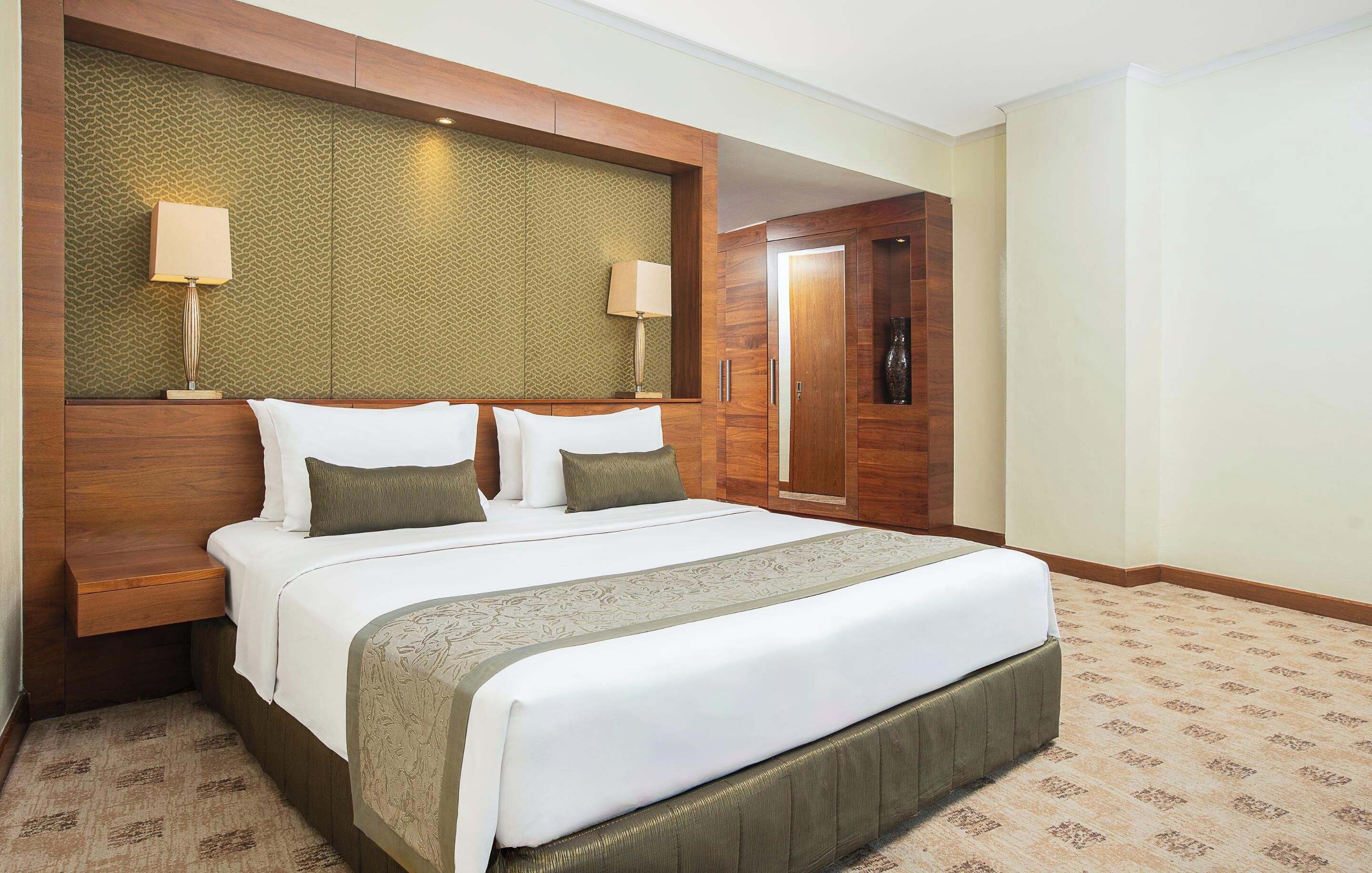 A hotel room with a large bed, wooden headboard, two bedside lamps, and a door leading to another room.