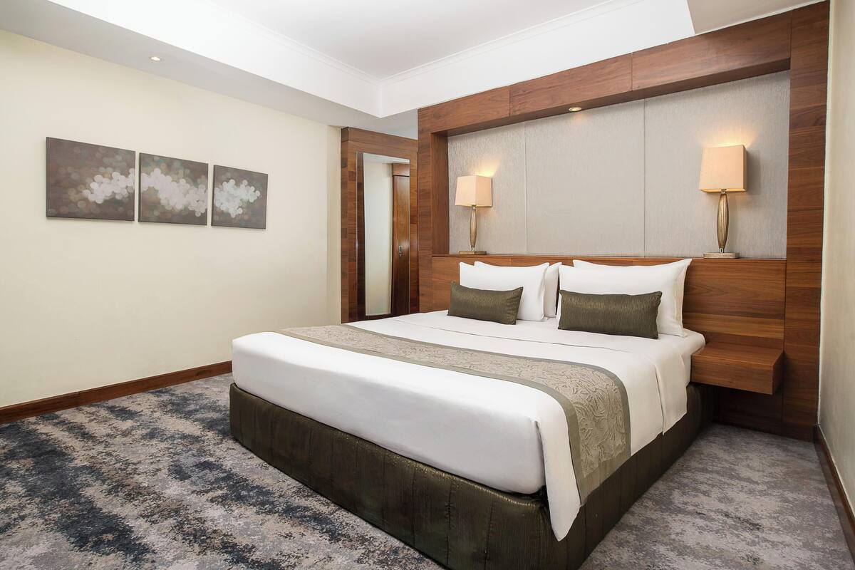 A hotel room with a large bed, wooden headboard, two bedside lamps, and a carpeted floor.