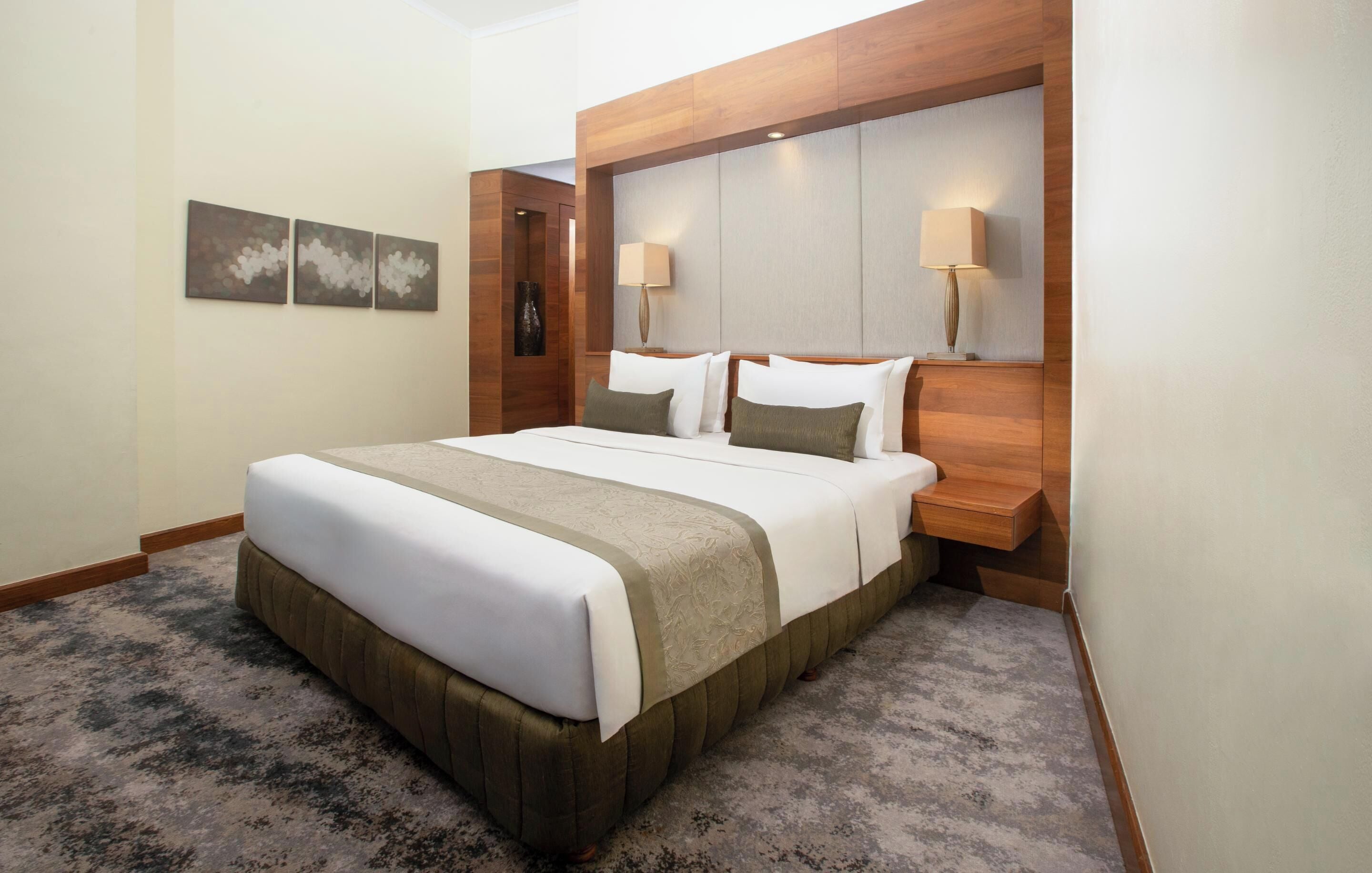 A hotel room with a large bed, wooden headboard, two bedside lamps, and a carpeted floor.