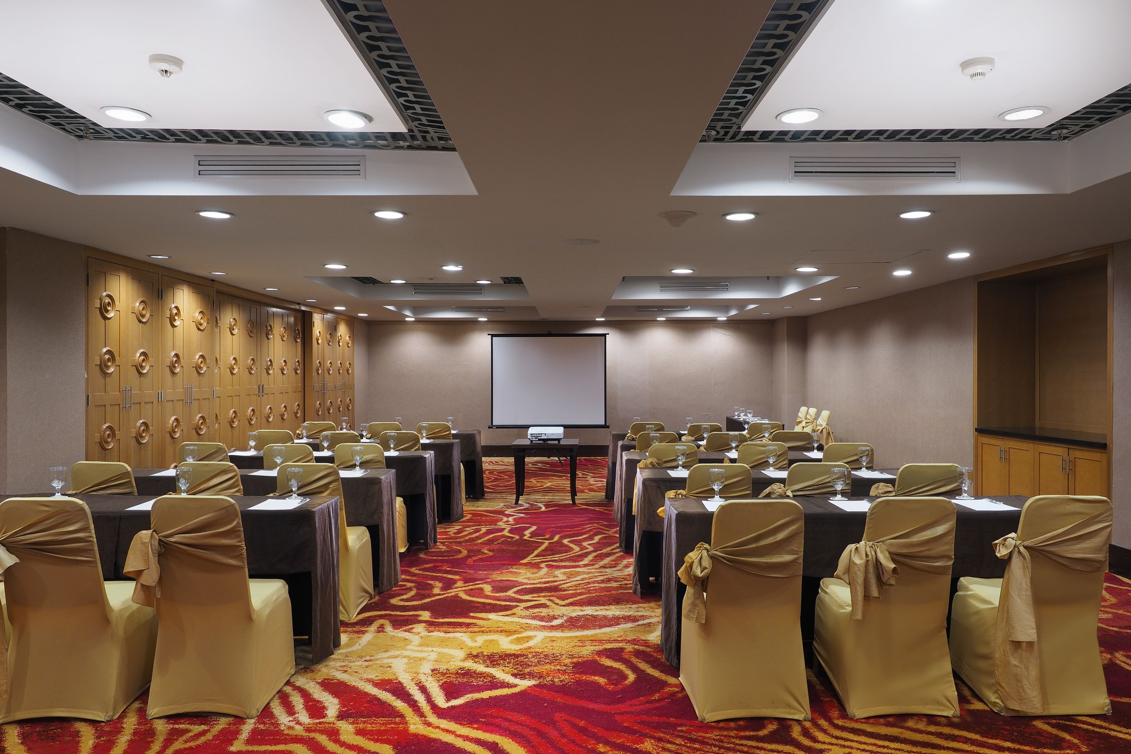 A conference room with rows of tables and chairs, a projector screen, and a carpeted floor.