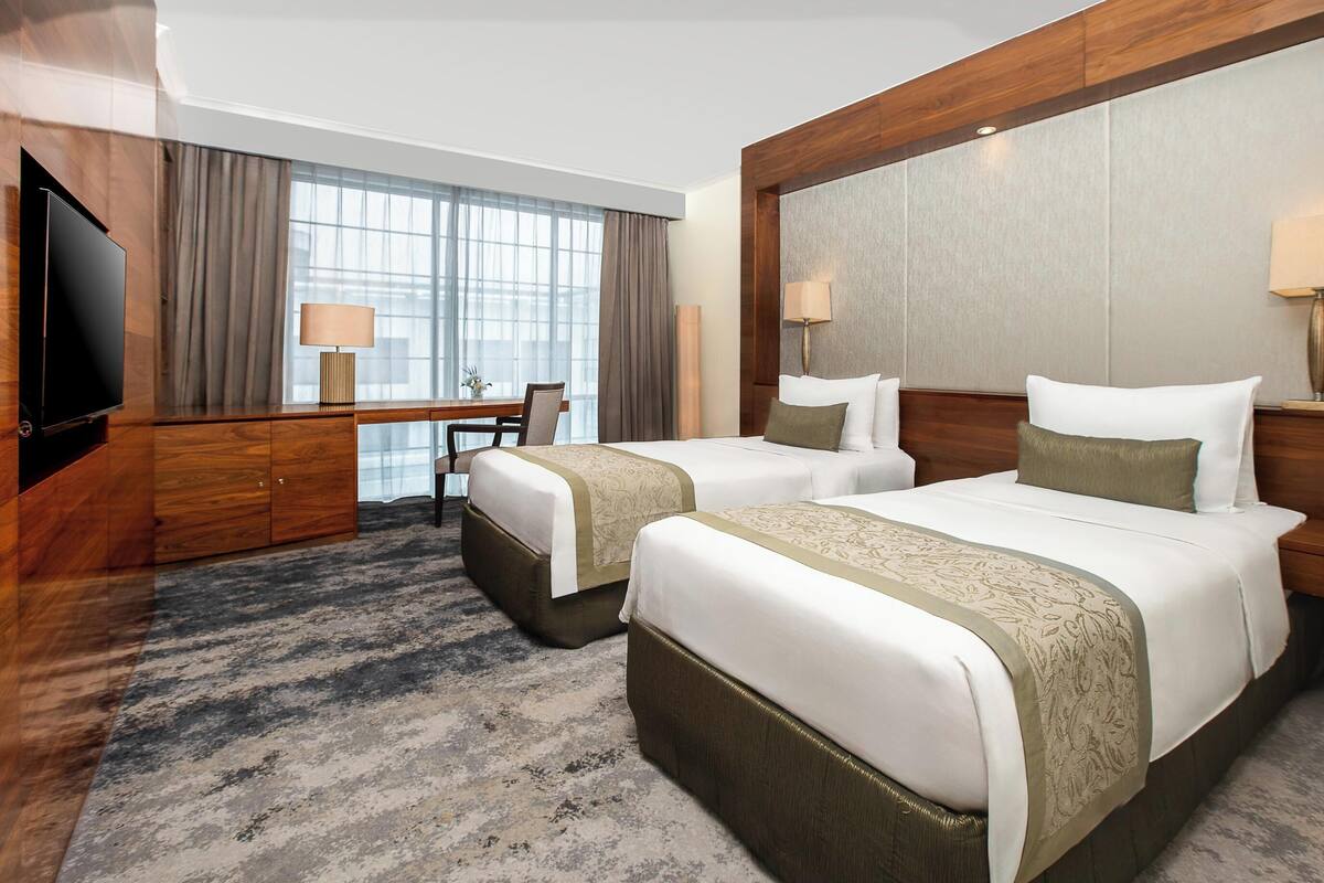 A hotel room with two beds, a desk, a television, and large windows with curtains.