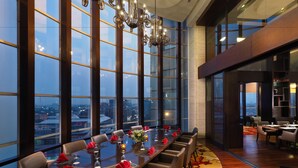 A dining area with a long table set for a meal, large windows offering a city view, and a modern interior design. - Aryaduta Medan (Medan)