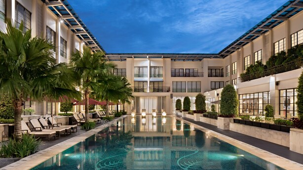 A modern indoor pool with clear blue water, surrounded by lounge chairs and palm trees, and a multi-story building with balconies and large windows. - Aryaduta Medan (Medan)