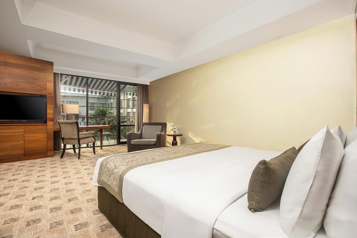 A hotel room with a large bed, a desk with a chair, a television, and a balcony with a view of buildings.