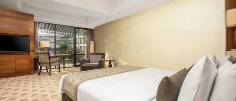 A hotel room with a large bed, a desk with a chair, a television, and a balcony with a view of buildings.