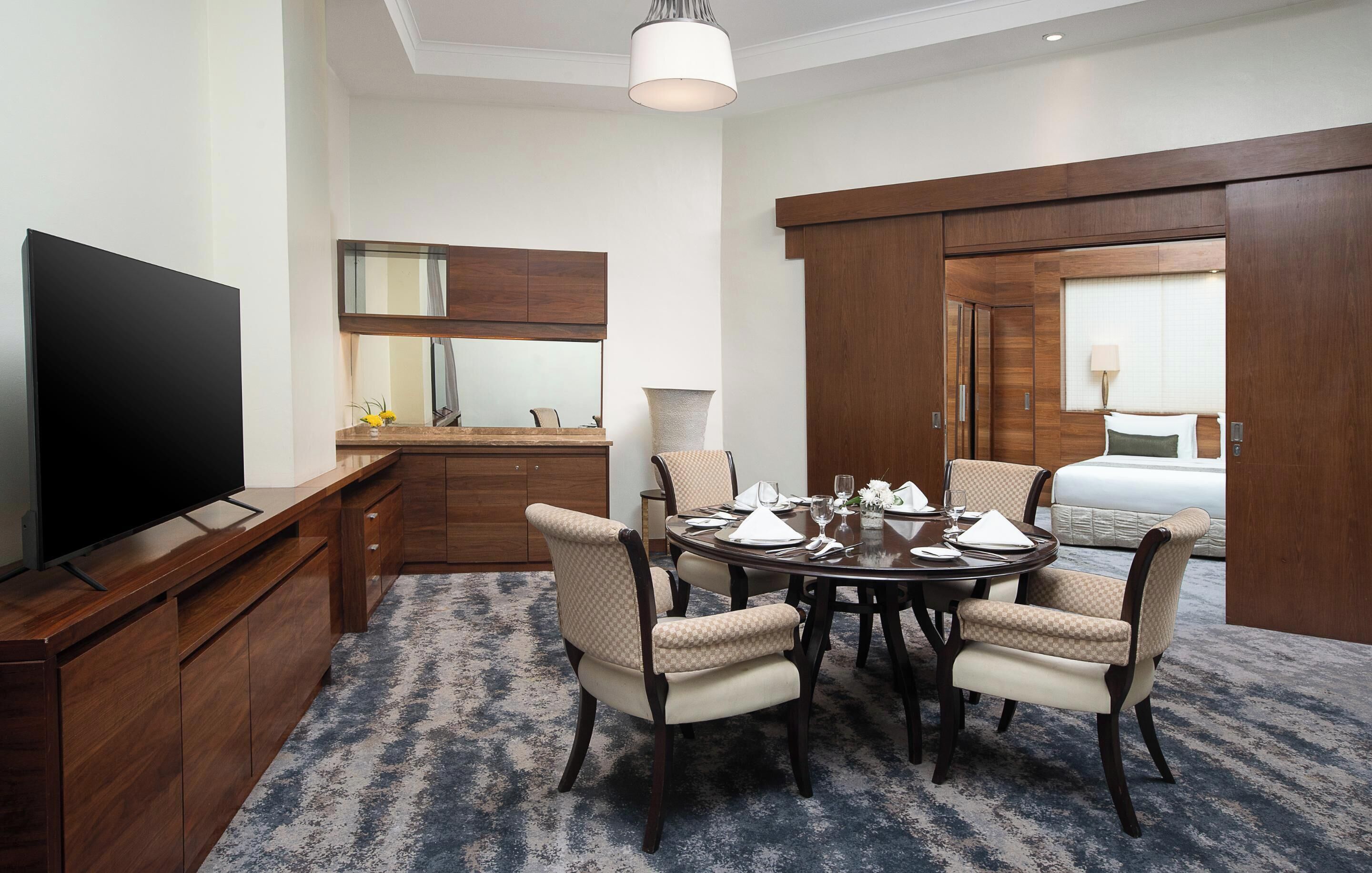 A hotel room with a TV, a dining table set for four, a large mirror, a wardrobe, and a bed.