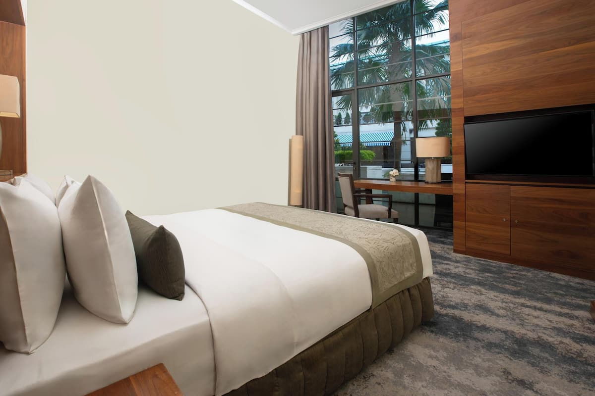 A hotel room with a large bed, a TV, a desk, and a window with a view of palm trees.