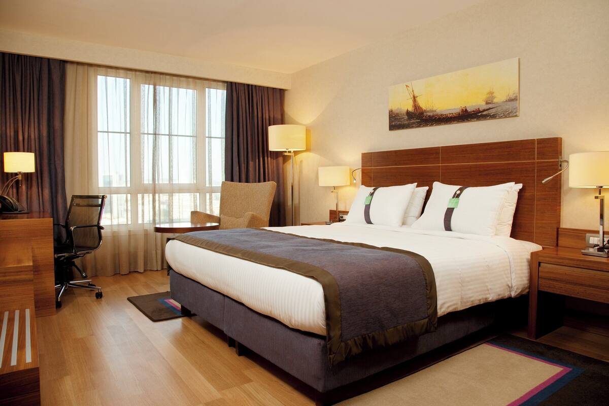 A hotel room with a large bed, two bedside tables with lamps, a desk with a chair, a window with curtains, and a painting on the wall.