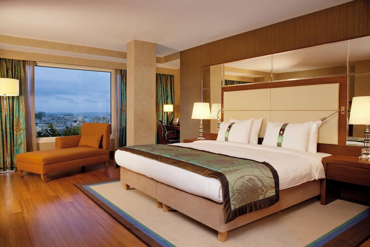 A hotel room with a large bed, a sofa, a nightstand, and a view of the city.