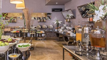 Montaje de bufé con diferentes estaciones de comida, que incluyen un bol de frutas, ensalada y varios dispensadores de bebidas.