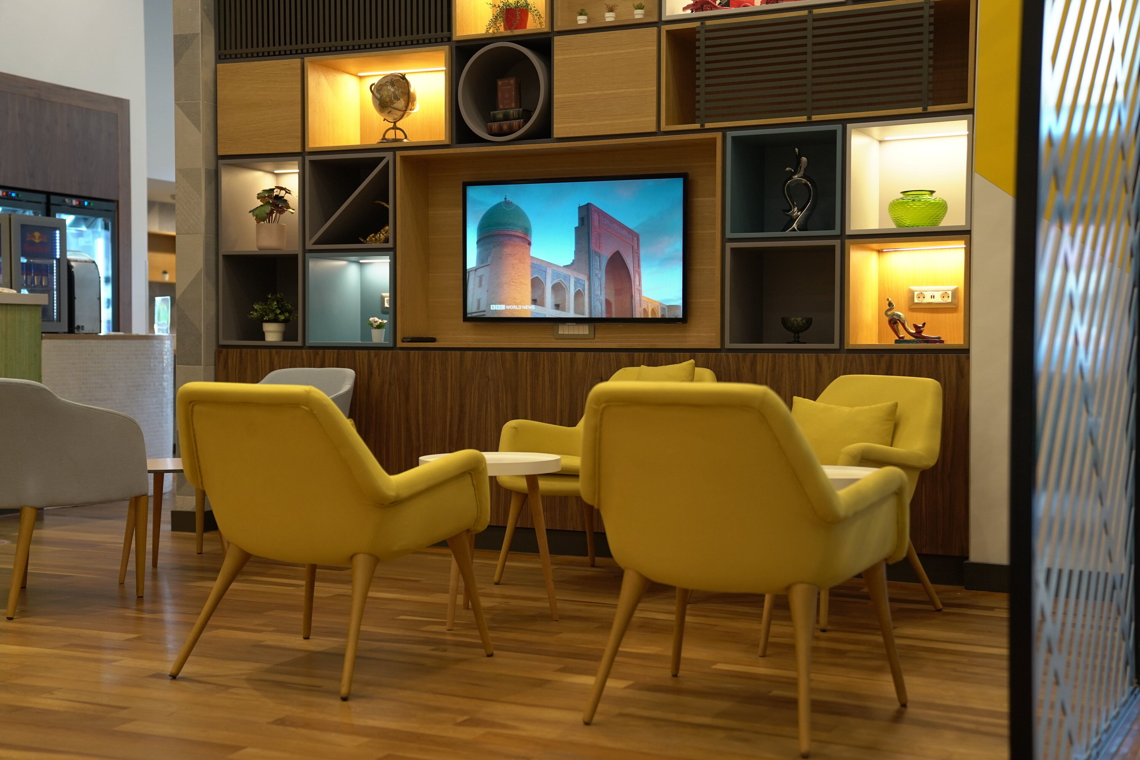 A modern interior with a TV displaying a building, wooden shelves, and yellow chairs.