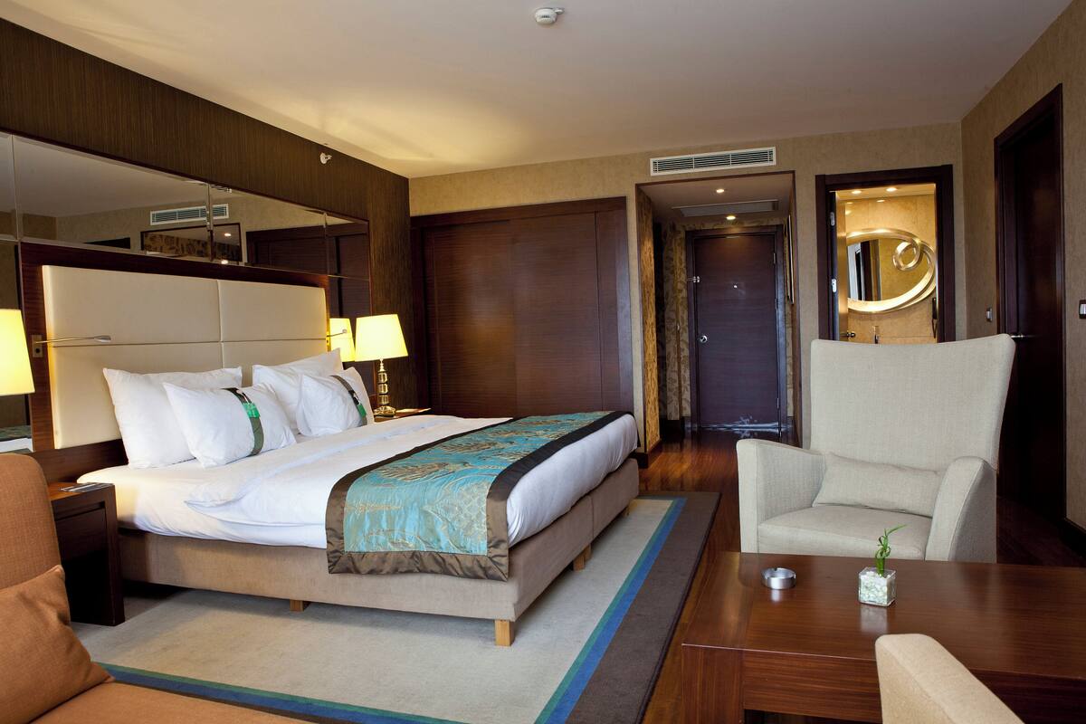A hotel room with a large bed, two bedside tables with lamps, a wooden desk, a chair, and a small plant.
