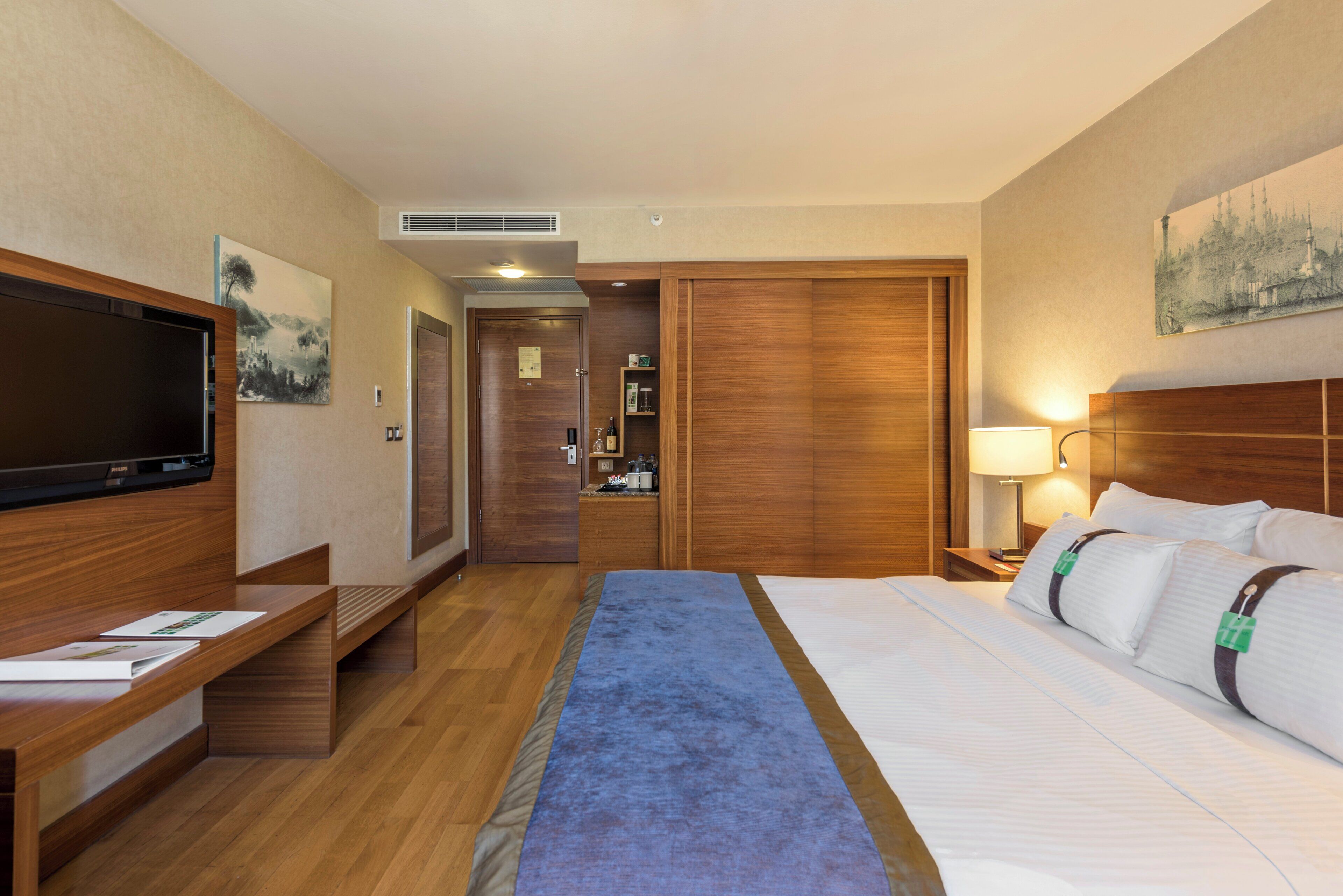 A hotel room with a large bed, a TV, a desk with papers, a nightstand with a lamp, and a wardrobe.