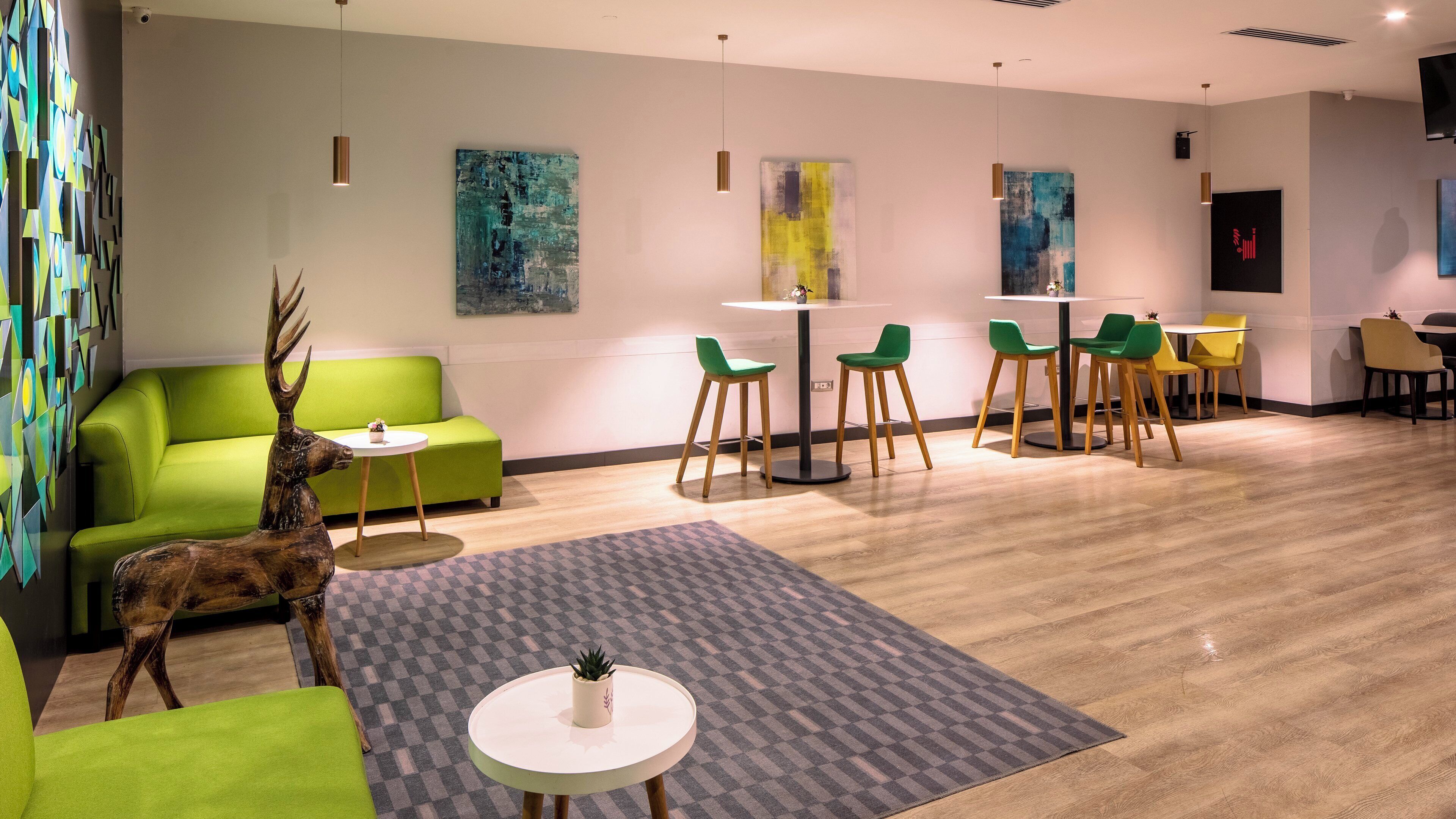 A modern hotel lobby with a green sofa, a small round table, and a decorative deer statue. There are multiple chairs and tables arranged for guests.