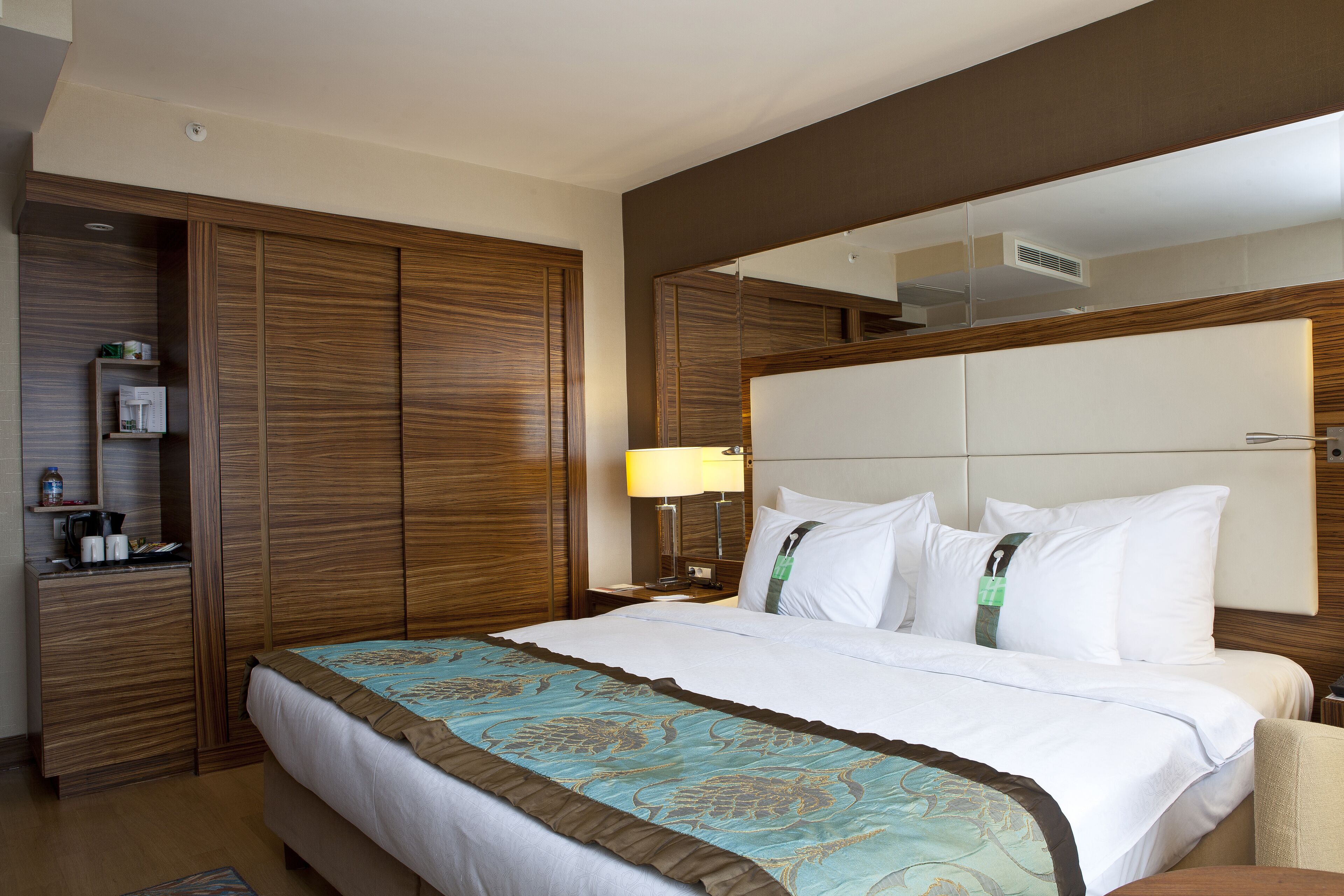 A hotel room with a large bed, wooden wardrobe, bedside table with a lamp, and a mirror above the bed.