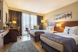 Une chambre d’hôtel avec deux lits, un bureau, une chaise, une lampe de bureau, un tableau au mur et une fenêtre avec des rideaux.