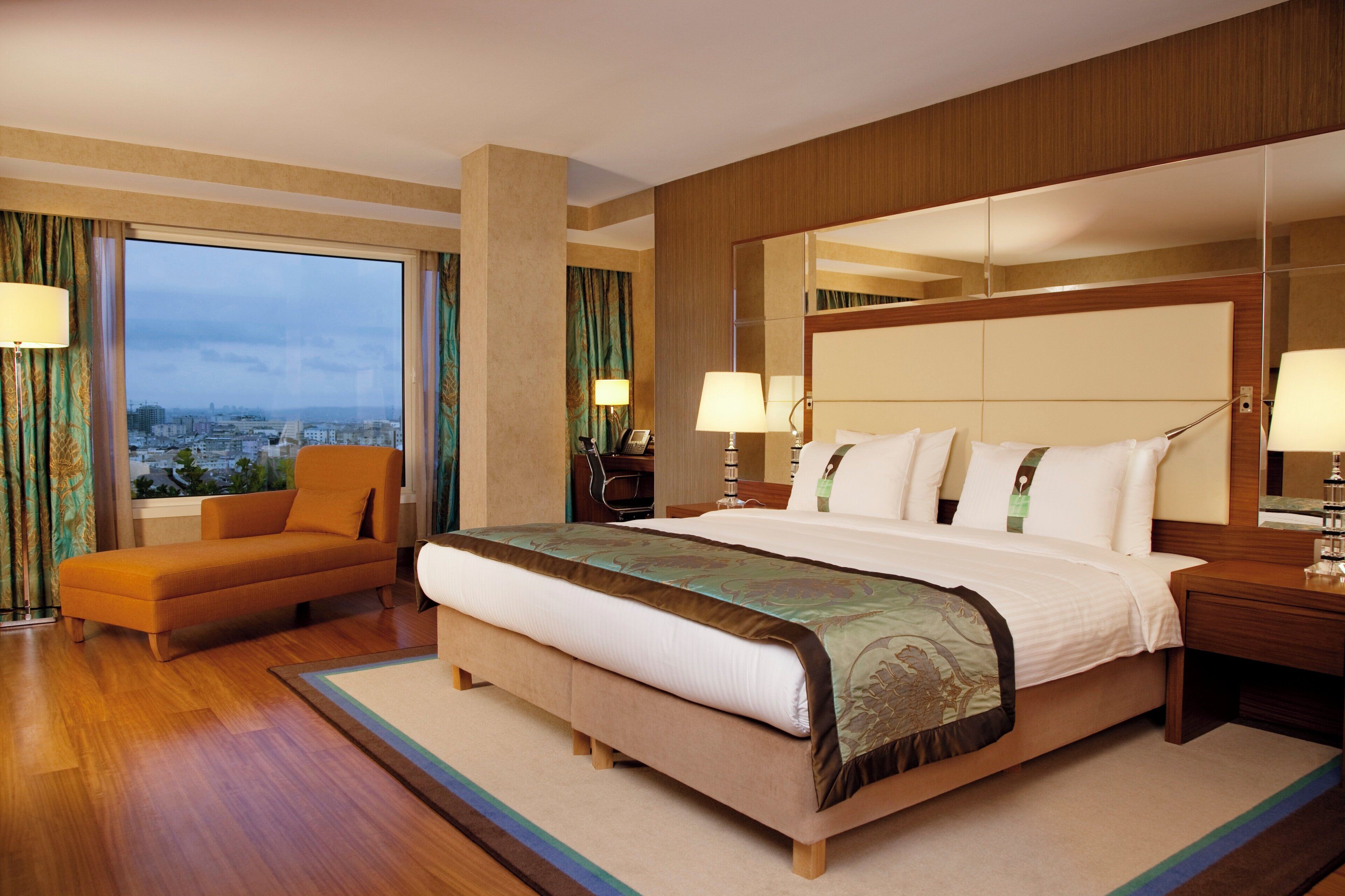 A hotel room with a large bed, a sofa, a nightstand, and a view of the city.