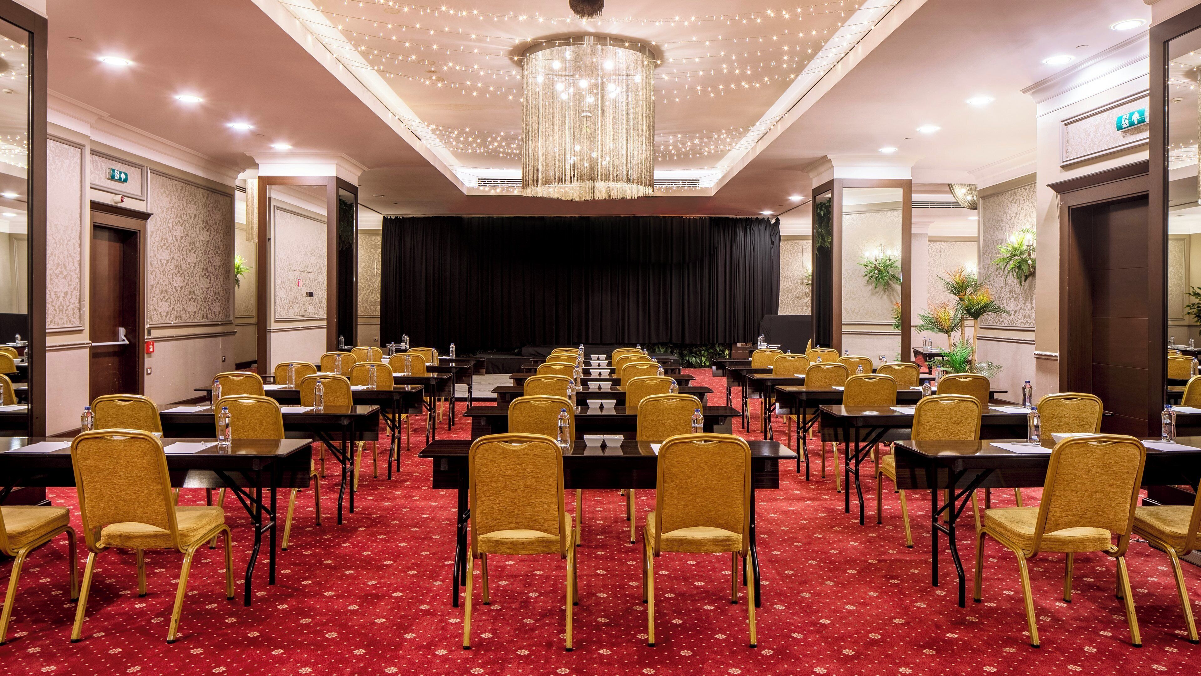 Une salle de conférence avec des rangées de tables et de chaises, une scène et un lustre.
