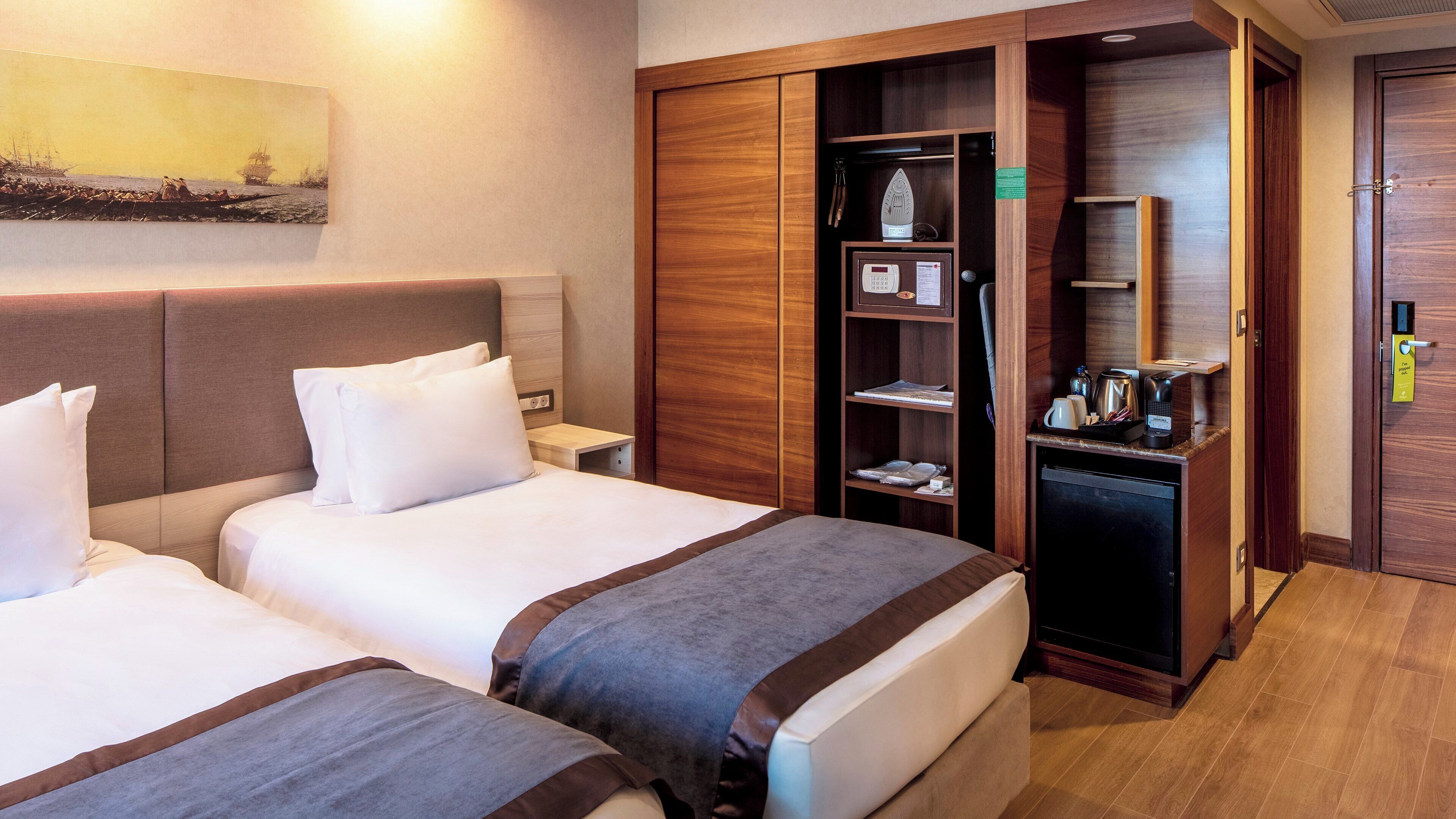 A hotel room with two beds, a wardrobe, a desk with a kettle, and a painting on the wall.