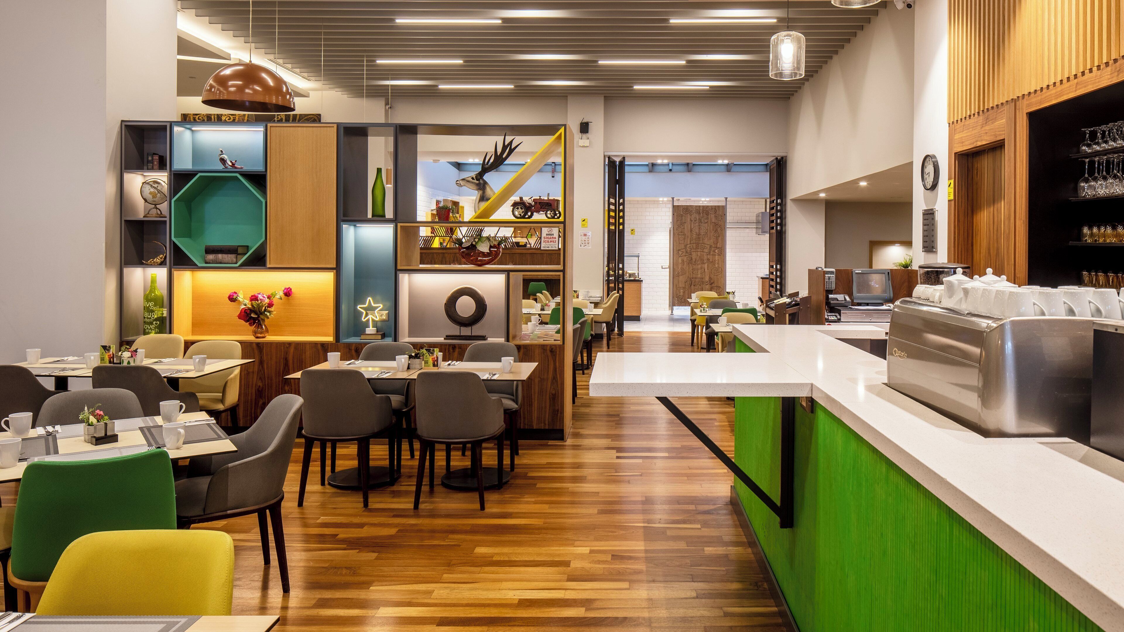 A modern restaurant with wooden flooring, green and yellow chairs, and a counter with a coffee machine.