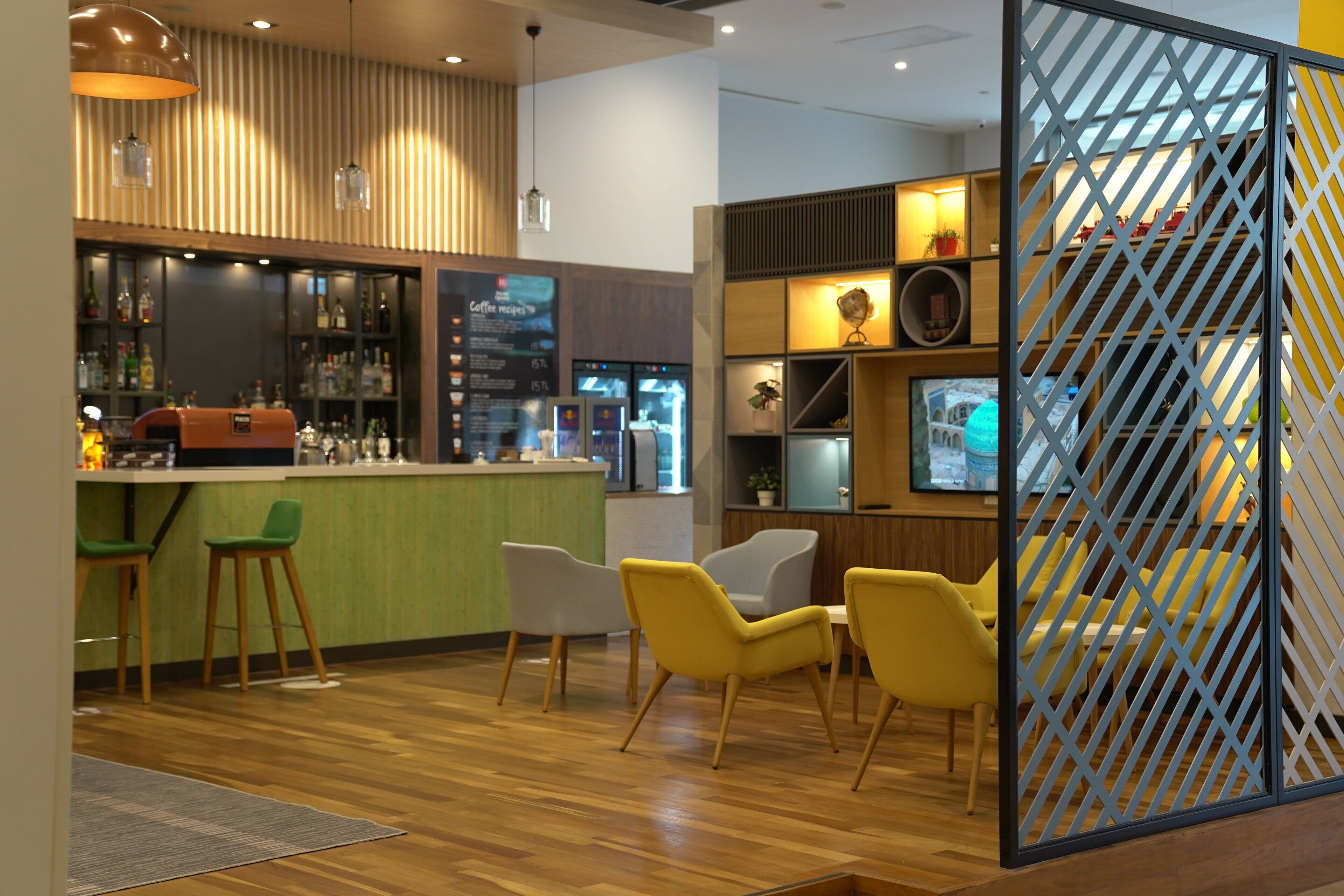 Un espace bar moderne avec un plancher en bois, des tabourets de bar verts et des chaises jaunes. On y trouve des étagères remplies de divers objets et un panneau de séparation orné d’un motif géométrique.