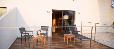 Una terraza de madera con dos sillas y una mesa, una puerta de vidrio y una pared blanca con un ventanal.