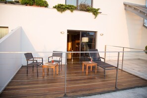 Une terrasse en bois avec deux chaises et une table, une porte d’entrée en verre et un mur blanc avec une fenêtre.