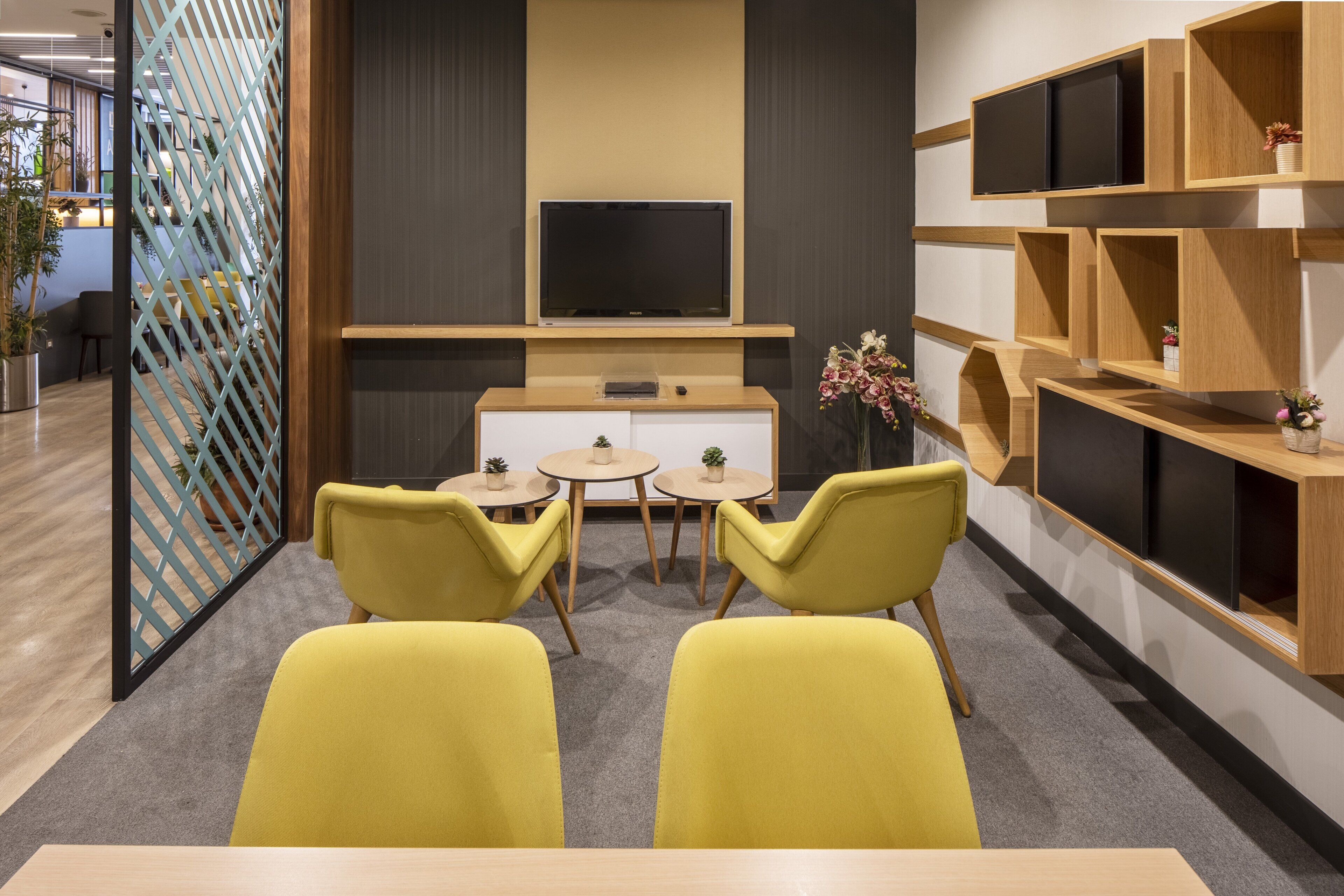A modern office space with two yellow chairs, a small round table, a TV on a wooden shelf, and a decorative plant.