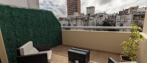 Una terraza en la azotea con mobiliario de mimbre, un seto verde y vistas a la ciudad.