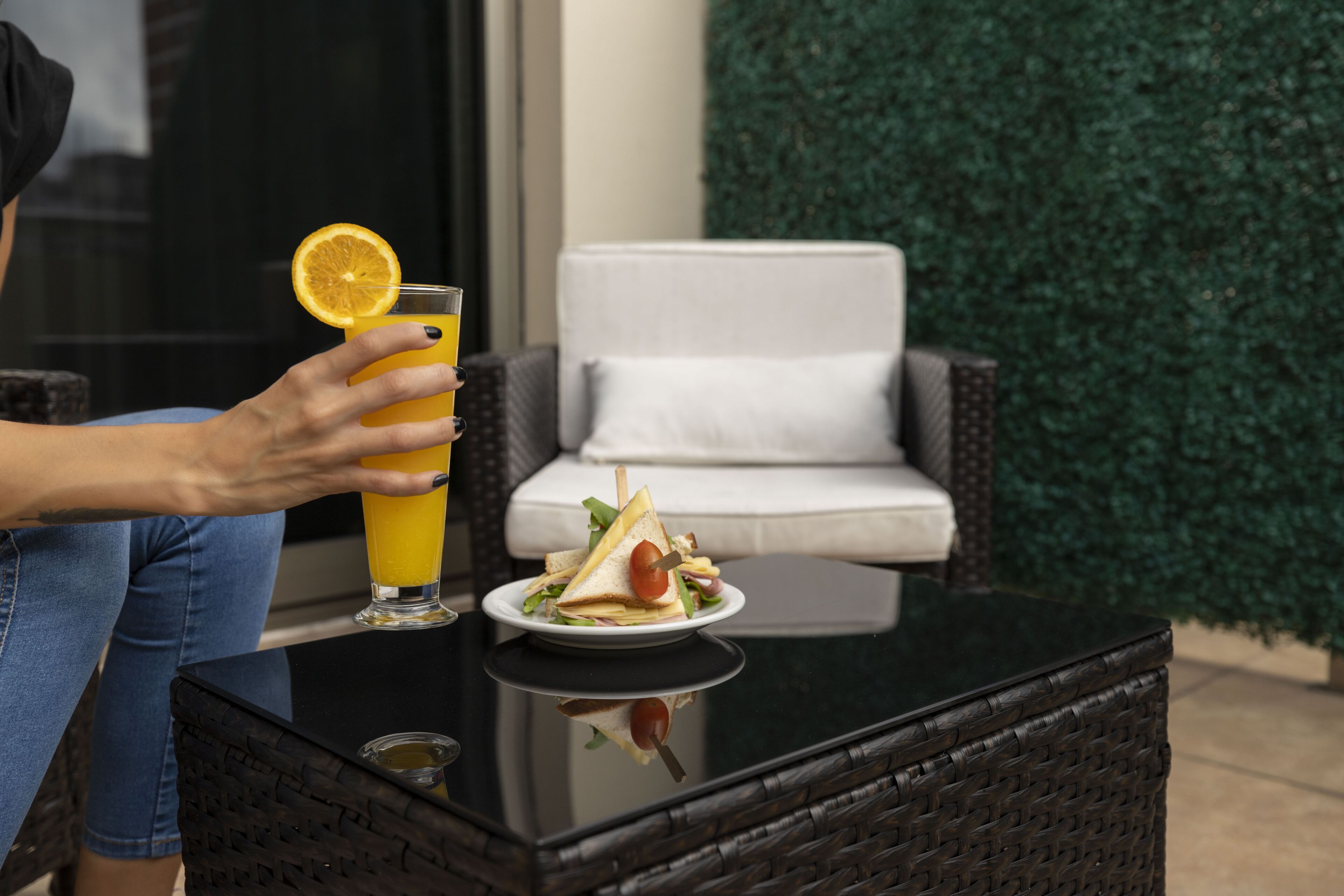 Eine Person hält ein Glas Orangensaft mit einer Zitronenscheibe und sitzt an einem Tisch mit einem Sandwich und Salat. Im Hintergrund sieht man einen Korbstuhl und eine grüne Hecke.