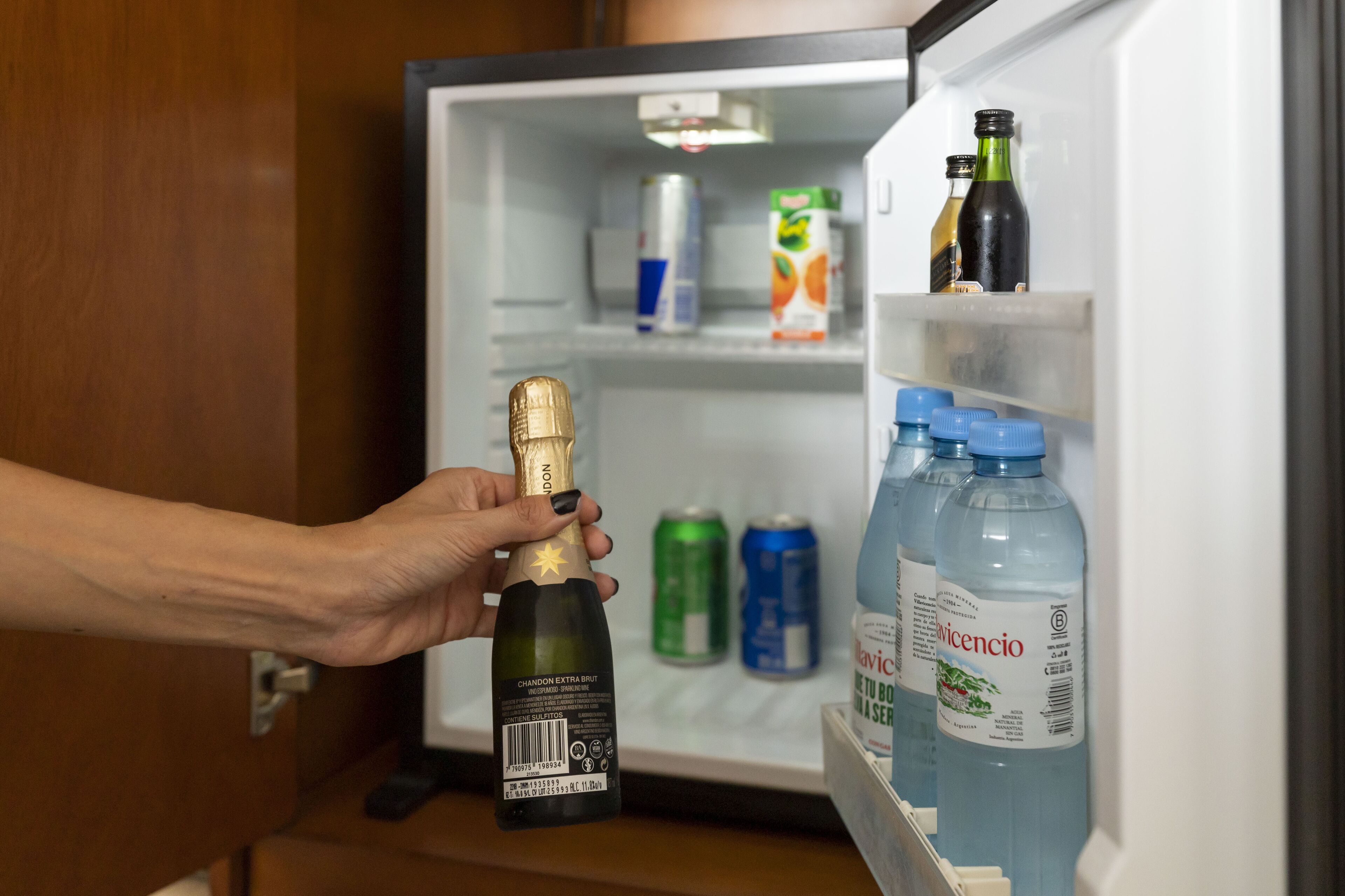 Una mano sostiene una botella de champán frente a un refrigerador abierto con diversas bebidas.