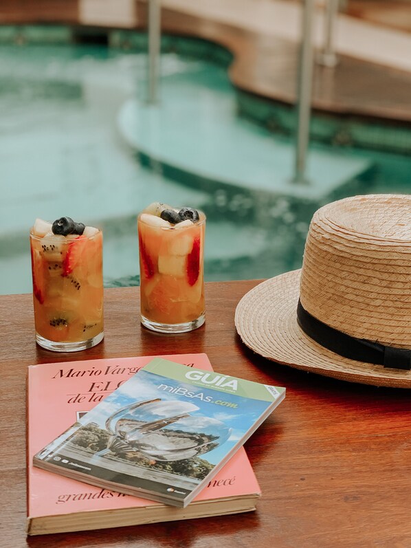 Two glasses of iced beverages with fruit on a wooden table, a straw hat, and a book with a travel guide cover.